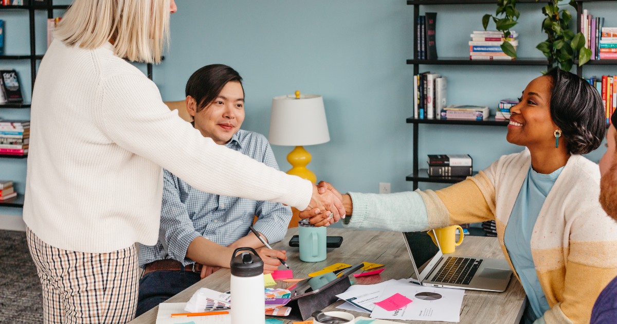 Two women shake hands while sitting with another person at a table