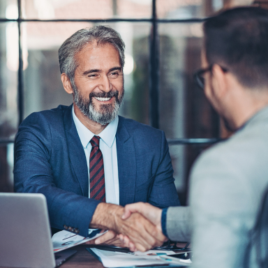 Two seated people shaking hands across a desk.