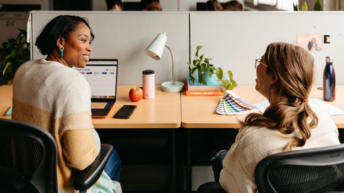 Two coworkers sitting at their side-by-side desks in conversation.