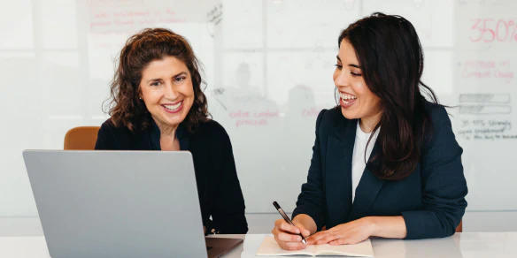 Two smiling women looking at laptop. One of the them is writing notes in paper notebook.