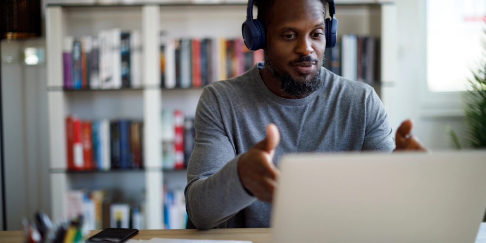 A man sitting at his desk wearing headphones and meeting on his laptop.